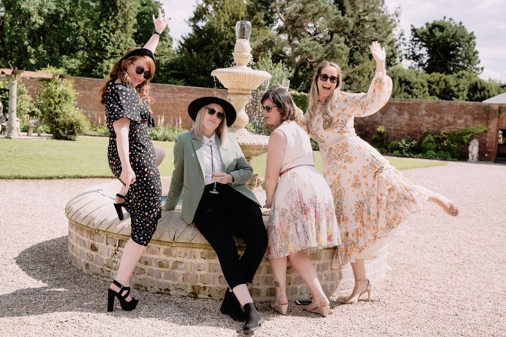 Four people pose playfully around a fountain in a garden at Elmhay Park, dressed in stylish summer outfits and sunglasses, capturing the essence of wedding photography with effortless charm.