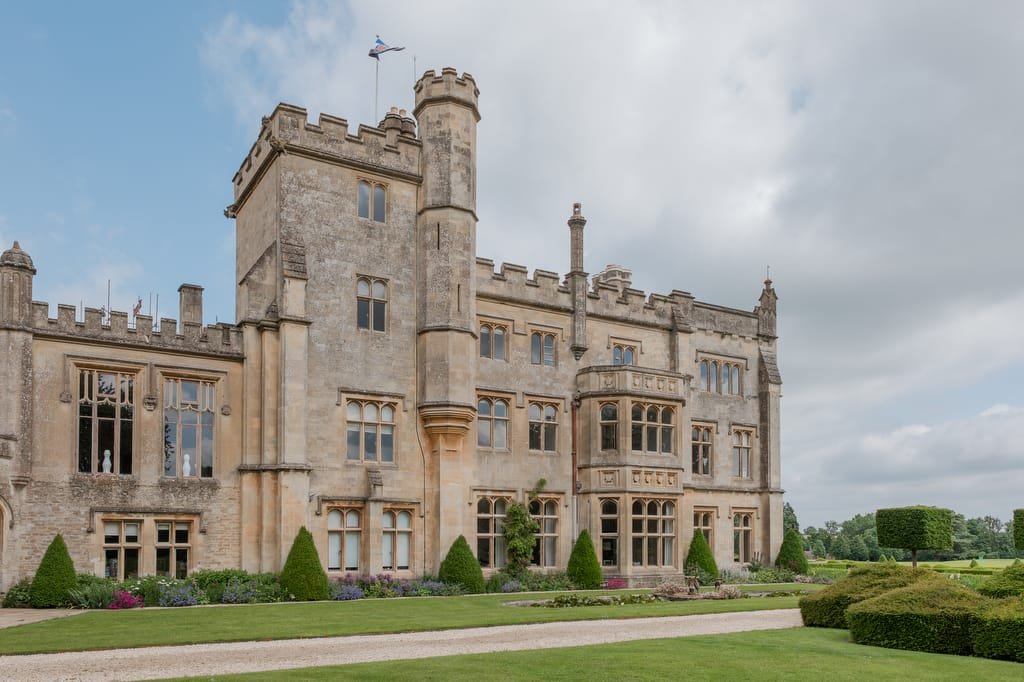 A large, stone castle-like mansion with a tower, surrounded by green lawns and gardens under a partly cloudy sky—a perfect setting captured elegantly by a Farleigh House wedding photographer.