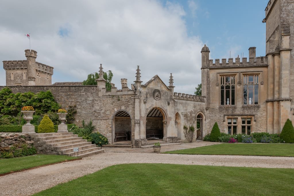 A historic stone building with a large arched entrance, set in a landscaped garden with manicured lawns, decorative urns, and an adjacent castle-like structure—an ideal scene for any Farleigh House Wedding Photographer.