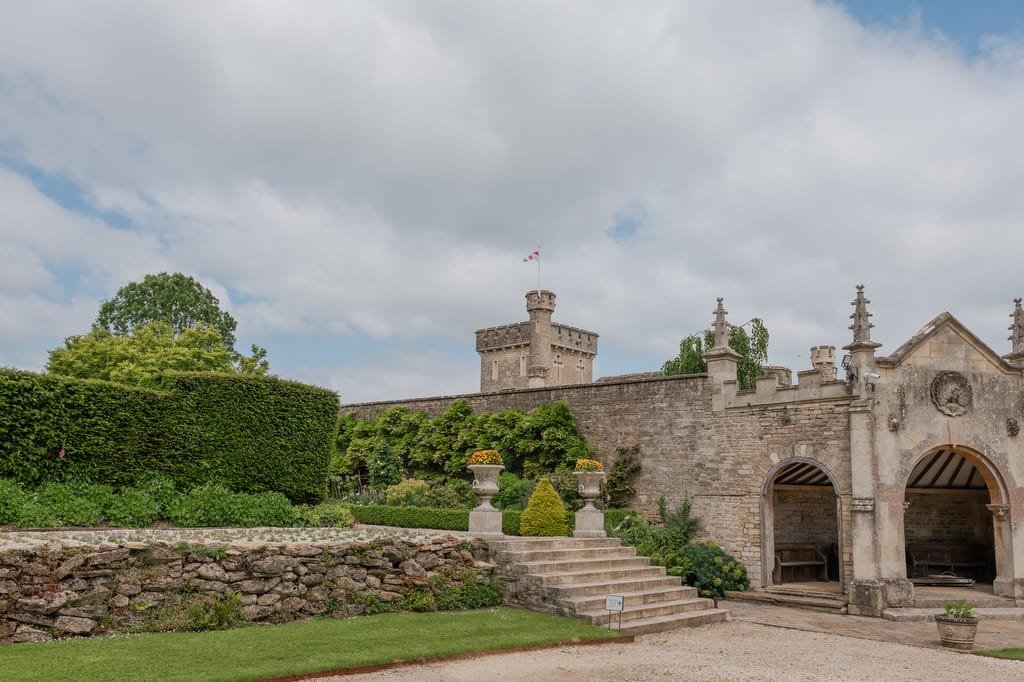 A historic stone building with ivy-covered walls, archways, and a central tower featuring a flag, set amidst a manicured garden under a partly cloudy sky—perfect scenery for any Farleigh House Wedding Photographer.