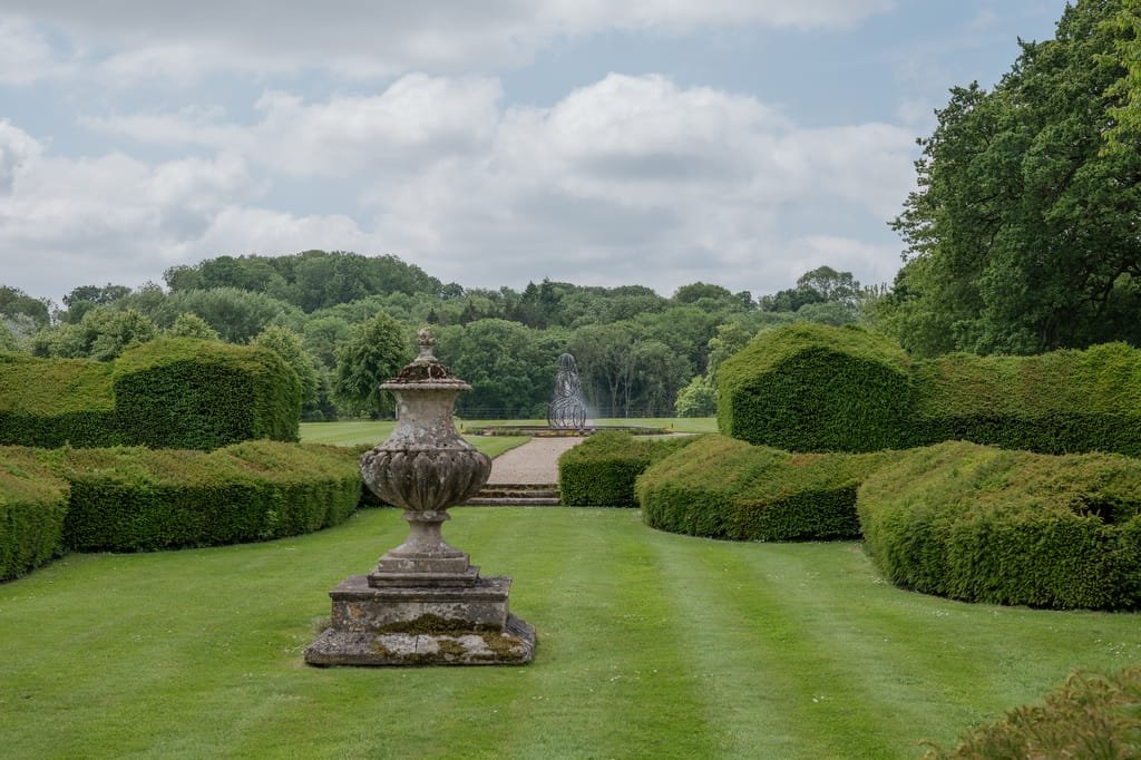 A formal garden at Farleigh House features manicured green lawns, neatly trimmed hedges, an ornate stone urn in the foreground, and a fountain in the distance. The background showcases a wooded area under a cloudy sky, providing an enchanting setting ideal for any wedding photographer.