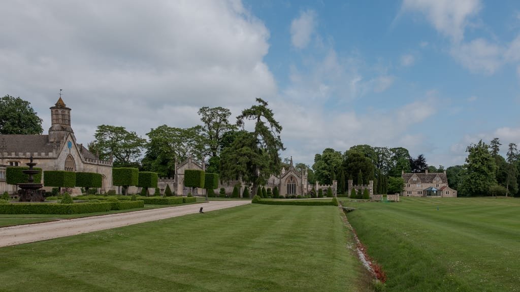 A large, historic estate under a cloudy blue sky, featuring manicured lawns, a stone path, a fountain, and multiple stone buildings surrounded by trees - an ideal backdrop for any Farleigh House Wedding Photographer.