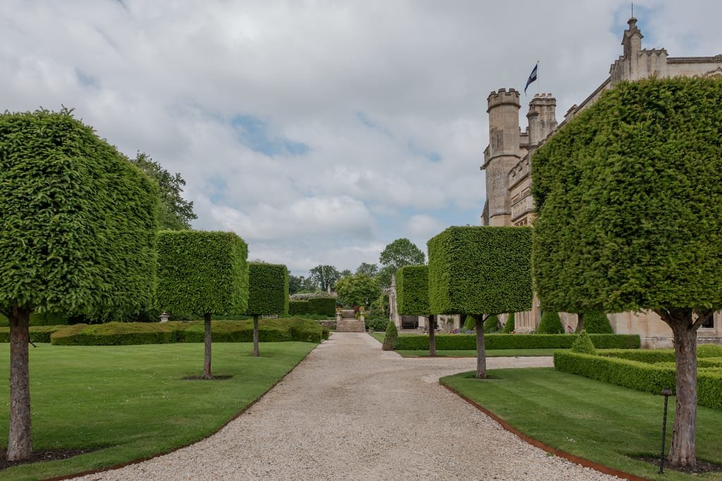 A gravel pathway lined with neatly trimmed rectangular trees leads to the historic stone building of Farleigh House. The sky is partly cloudy, and the manicured lawns are well-maintained, creating a picturesque setting for a wedding photographer.