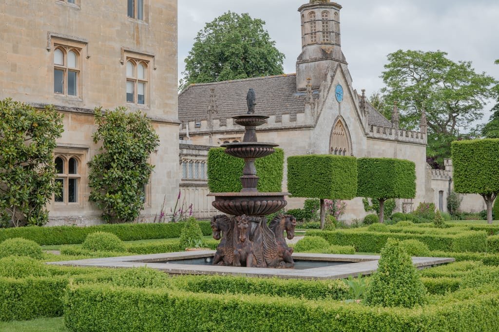 A historical stone building with neatly trimmed hedges and a decorative multi-tiered fountain featuring carvings of animal heads in the foreground, setting the perfect scene for a Farleigh House wedding photographer.