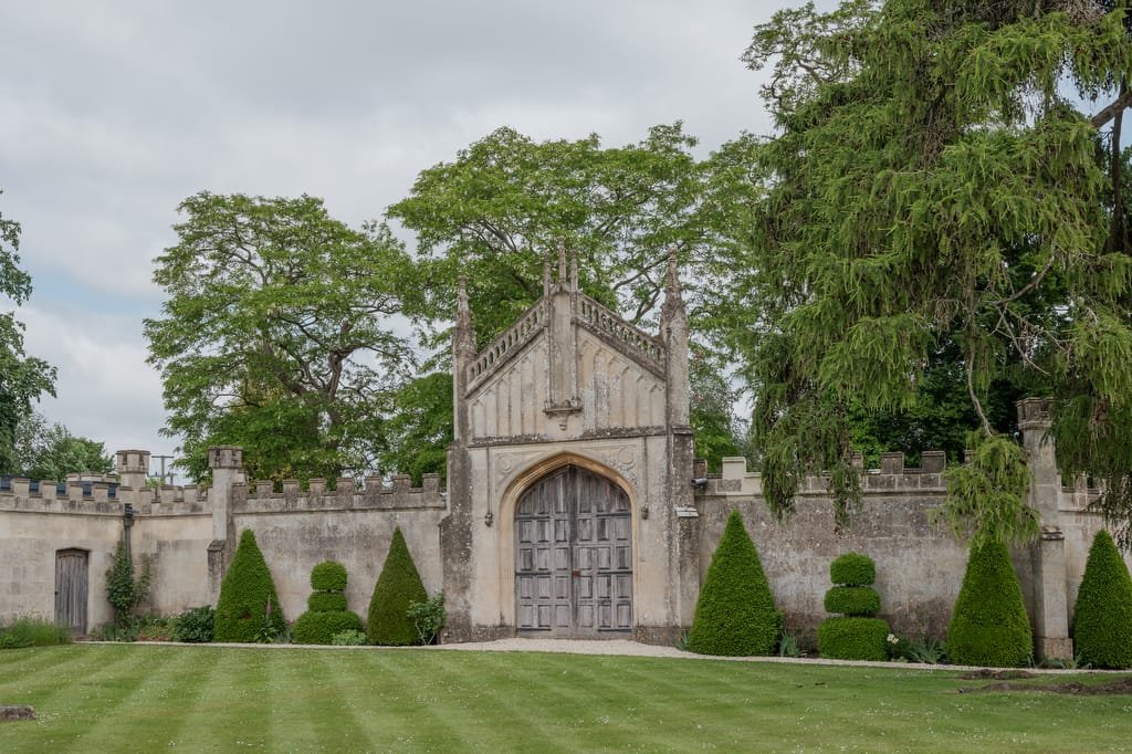 A large stone gate with a pointed arch and double wooden doors, flanked by greenery and neatly trimmed bushes in the foreground, with tall trees in the background—an enchanting scene perfect for capturing by a Farleigh House Wedding Photographer.