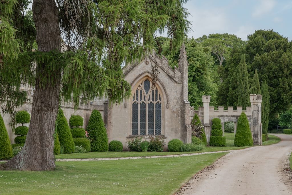 A stone chapel with a large arched window, surrounded by manicured shrubs and trees, sits adjacent to a dirt pathway leading through a well-maintained yard, perfect for capturing moments as a Farleigh House Wedding Photographer.