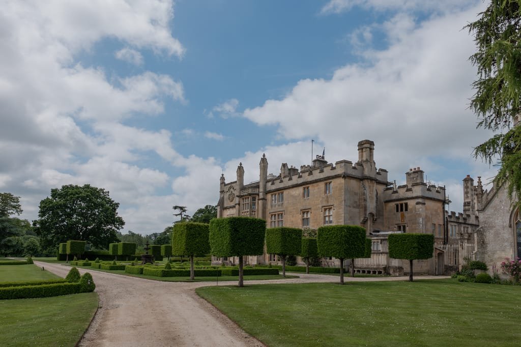 A large, historic stone building with ornate architecture, surrounded by manicured lawns, topiary trees, and a gravel path under a partly cloudy sky—a perfect setting for a Farleigh House wedding photographer to capture timeless moments.