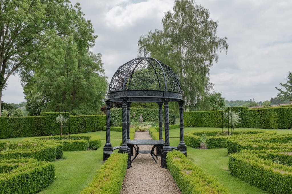 A black wrought iron gazebo with a domed roof stands on a gravel path, surrounded by manicured hedges and lush green trees in the formal gardens, creating an ideal backdrop for a Farleigh House wedding photographer.