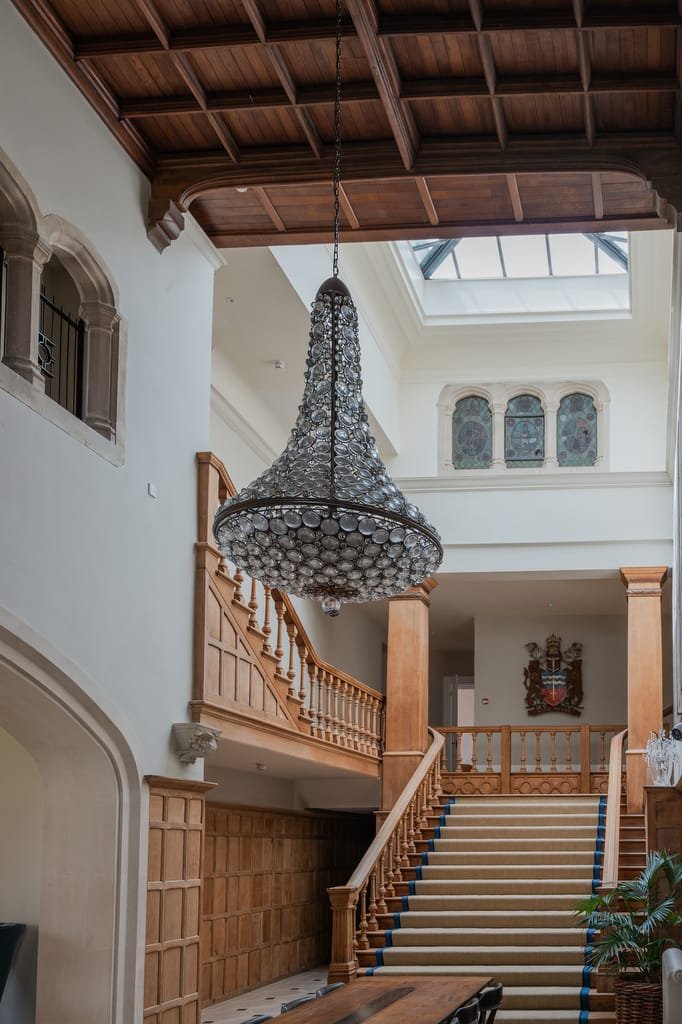 A grand interior space, perfect for a Farleigh House wedding photographer, featuring a large crystal chandelier hanging from a wooden ceiling, a staircase with wooden railings, and a coat of arms on the wall.