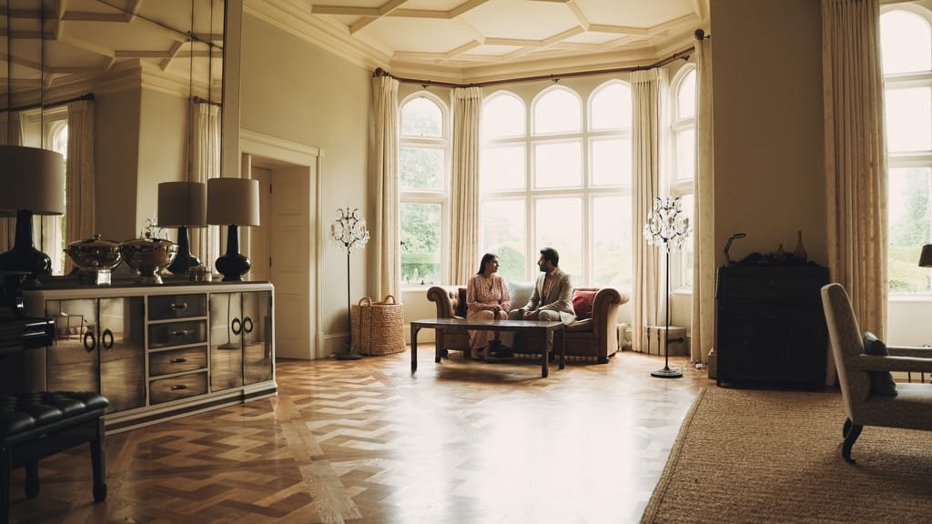 Two people sit on a sofa in a spacious, elegantly decorated living room with large windows and wooden flooring, the ambiance capturing the warmth and charm one might find in a Farleigh House Wedding Photographer's portfolio.