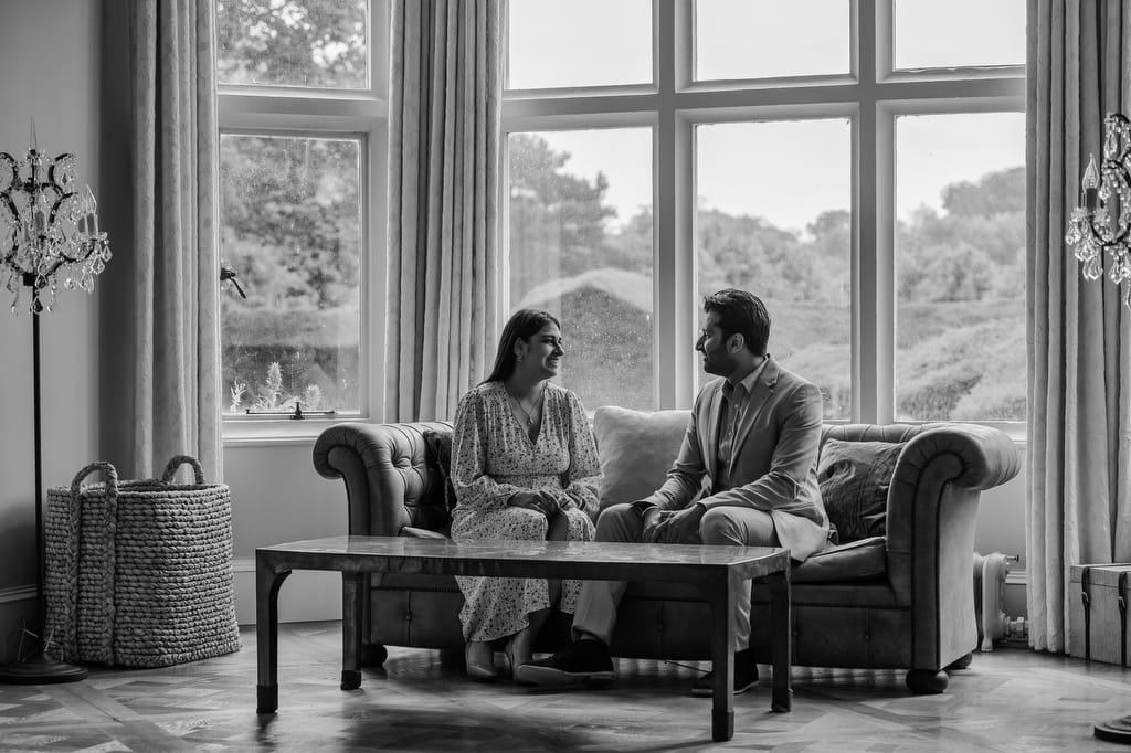 A woman and a man sit on a couch facing each other in front of large windows, engaging in conversation in a living room. The black-and-white image, reminiscent of a Farleigh House Wedding Photographer’s work, showcases a calm and well-lit interior space.