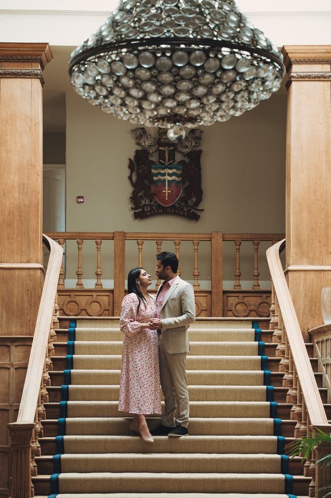 A couple stands facing each other on a grand staircase with wooden railings, under a large chandelier, in a room adorned with a crest on the wall—a perfect scene for any Farleigh House Wedding Photographer to capture.