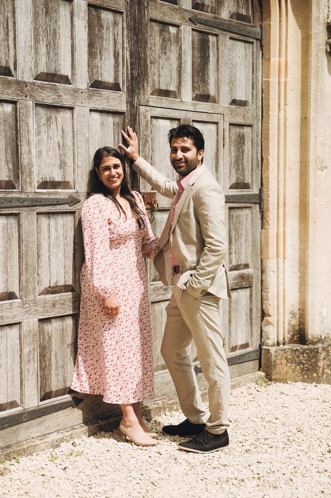 A man and woman stand in front of large wooden doors. The woman is wearing a floral dress, and the man is in a light-colored suit with a pink tie. They are both smiling and facing the camera, captured beautifully by their Farleigh House wedding photographer.
