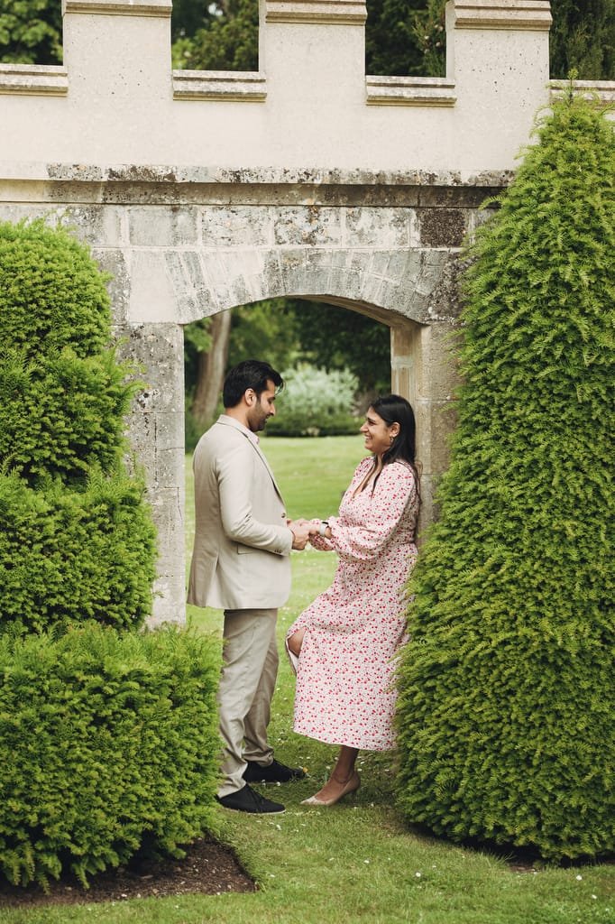 A man and woman stand facing each other, holding hands, in front of a stone archway with greenery around them. The man wears a beige suit, and the woman wears a pink floral dress—a perfect moment captured by a Farleigh House wedding photographer.