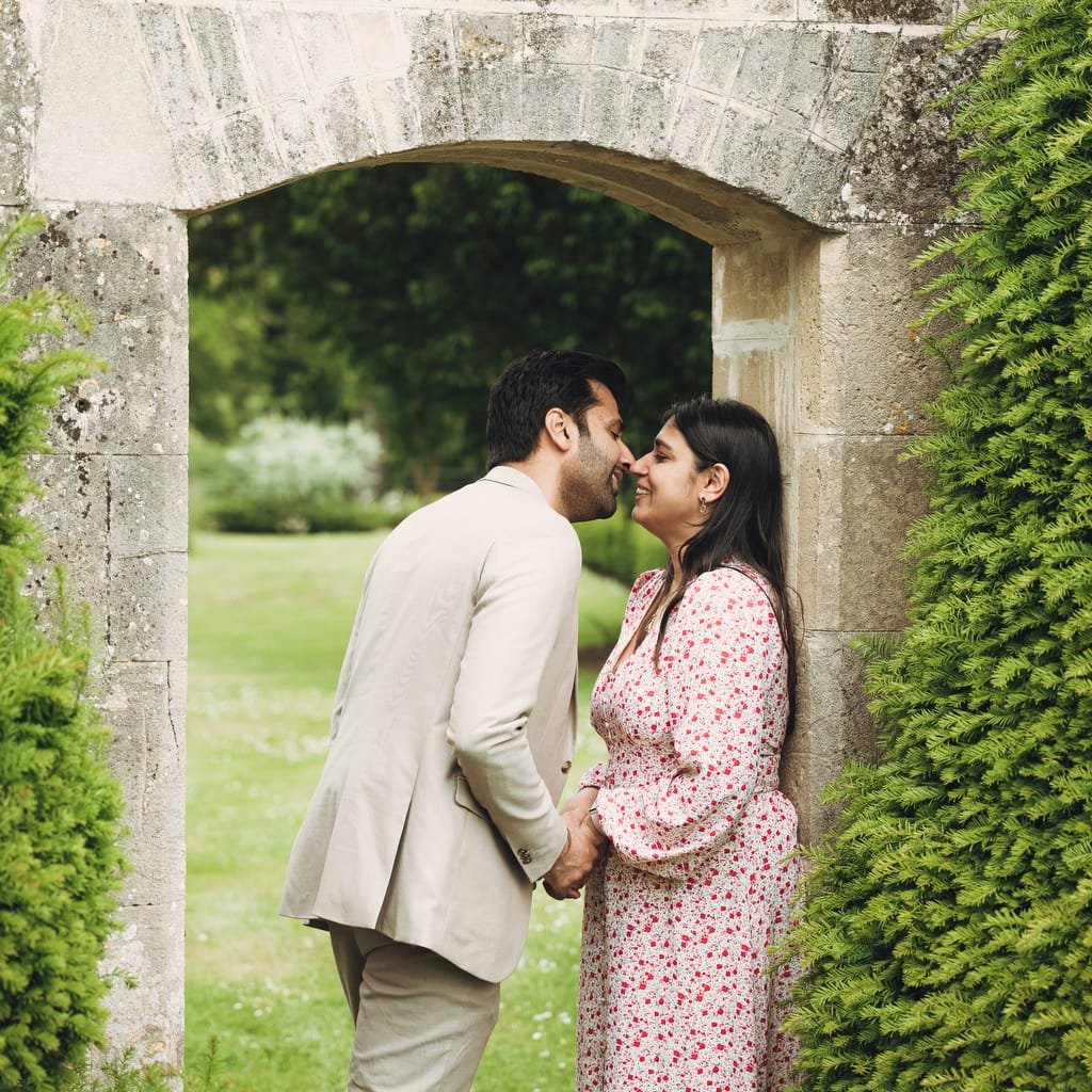 A man and woman stand under a stone archway, kissing and holding hands. They are surrounded by greenery, captured beautifully by a Farleigh House Wedding Photographer.