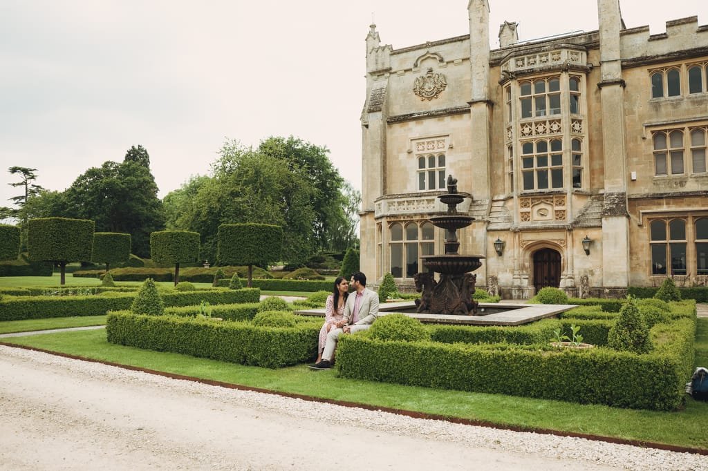 A couple sits on a bench near a fountain in front of a historic stone building surrounded by hedges and gardens, as captured beautifully by a Farleigh House wedding photographer.