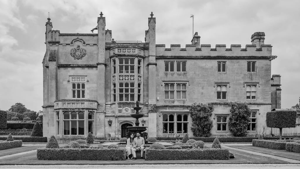 Black and white image of an old, grand building with intricate architecture. A group of people is sitting on a bench in the foreground, surrounded by neatly manicured gardens—an ideal setting for a Farleigh House Wedding Photographer.