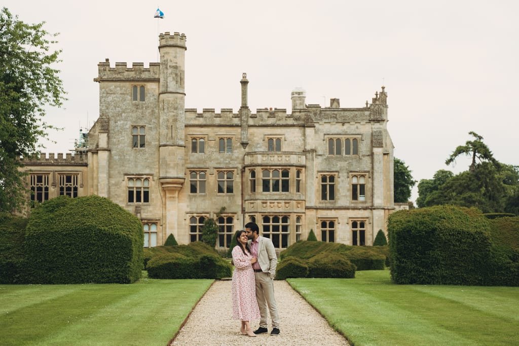 A couple stands close together on a gravel path, posing in front of the large, historic Farleigh House with manicured lawns and gardens, captured beautifully by their wedding photographer.