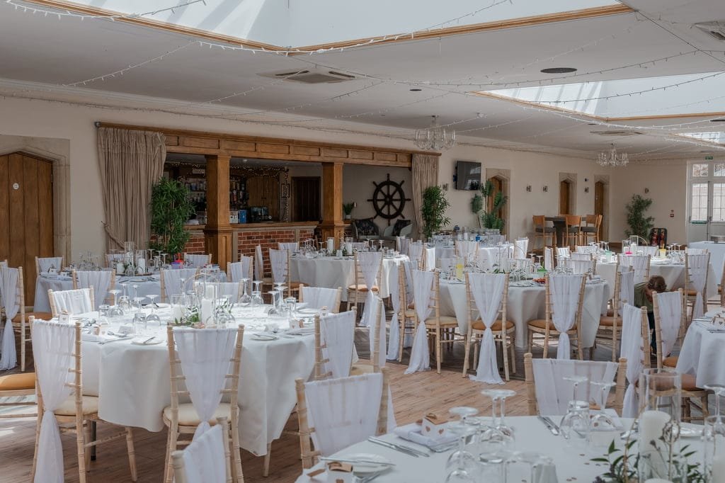 A banquet hall set up for an event at Elmhay Park, featuring round tables with white tablecloths and chairs with white covers, decorative plants, and a wooden bar area in the background—ideal for weddings or capturing moments as a photographer.