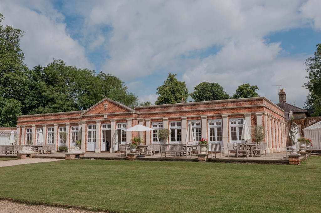 A single-story brick building with multiple French doors and windows, featuring outdoor seating areas with tables and umbrellas, set against a backdrop of green lawns and trees under a partly cloudy sky—perfect for capturing moments by a weddings photographer at Elmhay Park.