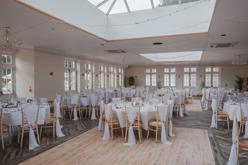 A large event hall at Elmhay Park with several round tables decorated with white tablecloths and chairs draped in white fabric, set up for a formal gathering. Overhead string lights create a charming ambiance, perfect for weddings. A photographer's dream setting.