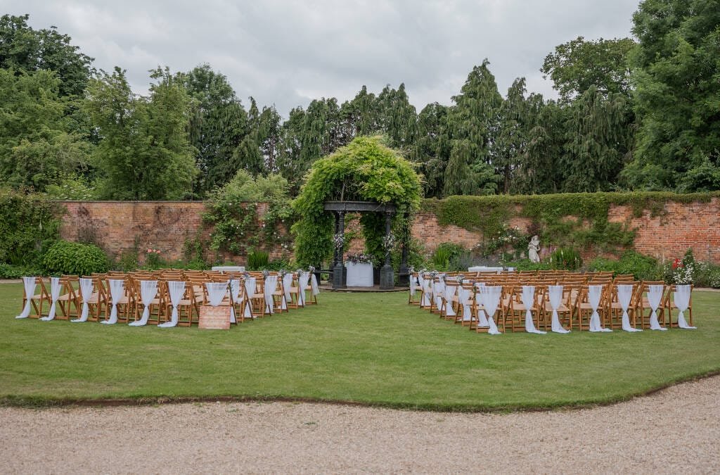 An outdoor wedding ceremony setup at Elmhay Park with rows of wooden chairs, white chair sashes, and an altar adorned with greenery and flowers, situated on a lawn area with a brick wall and trees in the background, perfect for capturing by a weddings photographer.