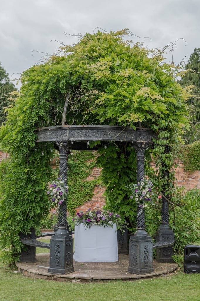 A metal gazebo draped with lush greenery and purple flowers, set in a garden perfect for weddings at Elmhay Park, featuring a circular table covered with a white cloth underneath—an ideal setting for any photographer's dream.