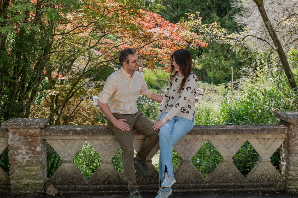 Two people sit on a stone railing in a garden, engaged in conversation. The background features vibrant green foliage and autumn-colored trees, reminiscent of the serene ambiance found at the Temple of Minerva Bath.