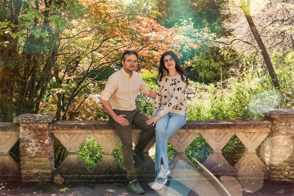 A man and a woman sit on a stone railing in the garden of the Temple of Minerva Bath, with sunlit trees and foliage in the background.