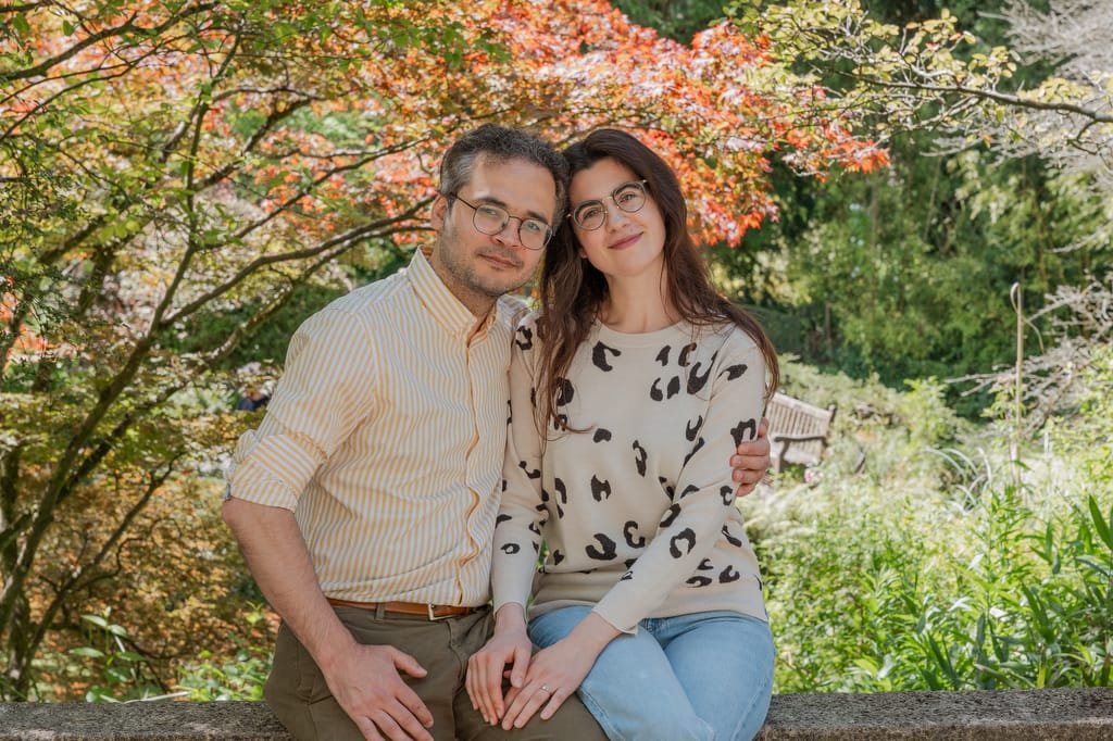 A man and a woman sit close together on a stone ledge in the garden at the Temple of Minerva Bath, surrounded by autumnal foliage.