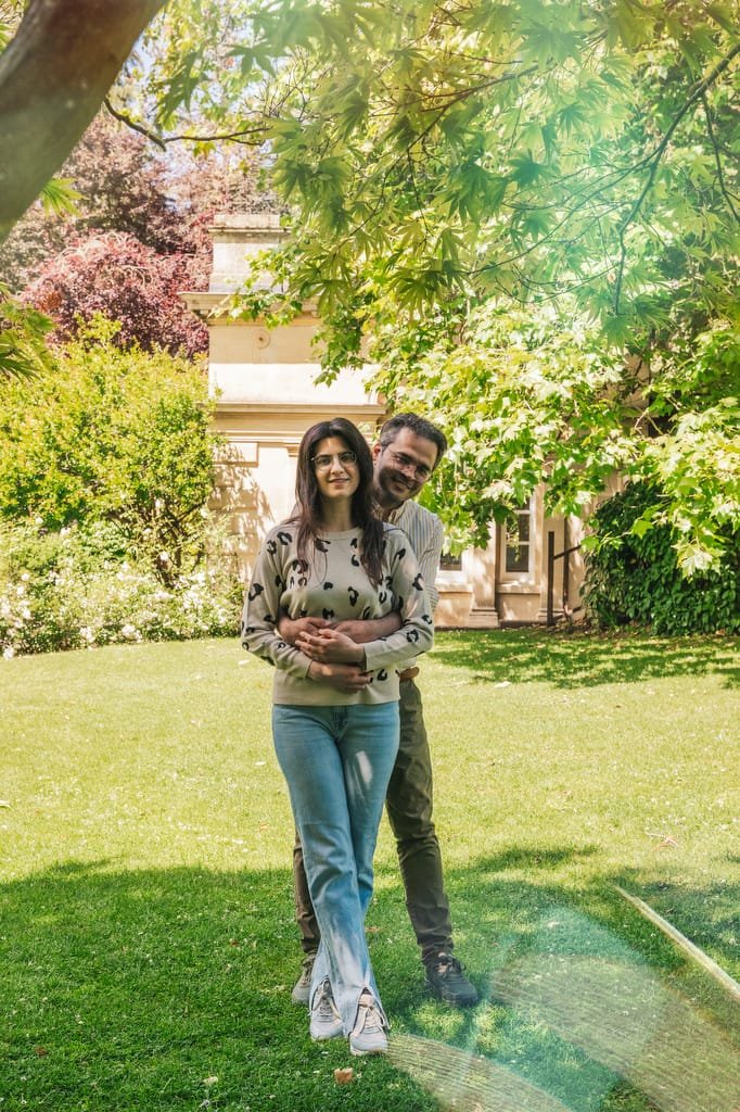 A man and woman standing in a sunny garden with green foliage near the Temple of Minerva Bath. The man is behind the woman with his arms around her waist. Both are smiling and casually dressed.