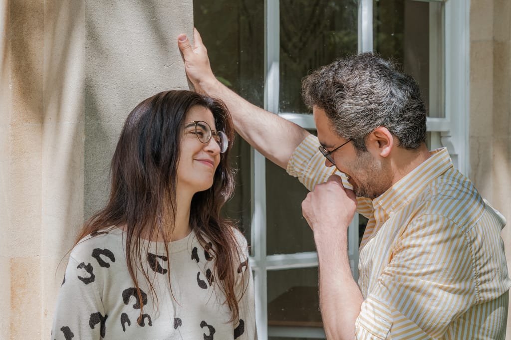 A woman with long hair and glasses smiles while a man in a striped shirt and glasses leans against a wall, looking at her. They stand outside near a large window, reminiscent of the grand architecture of the Temple of Minerva Bath.