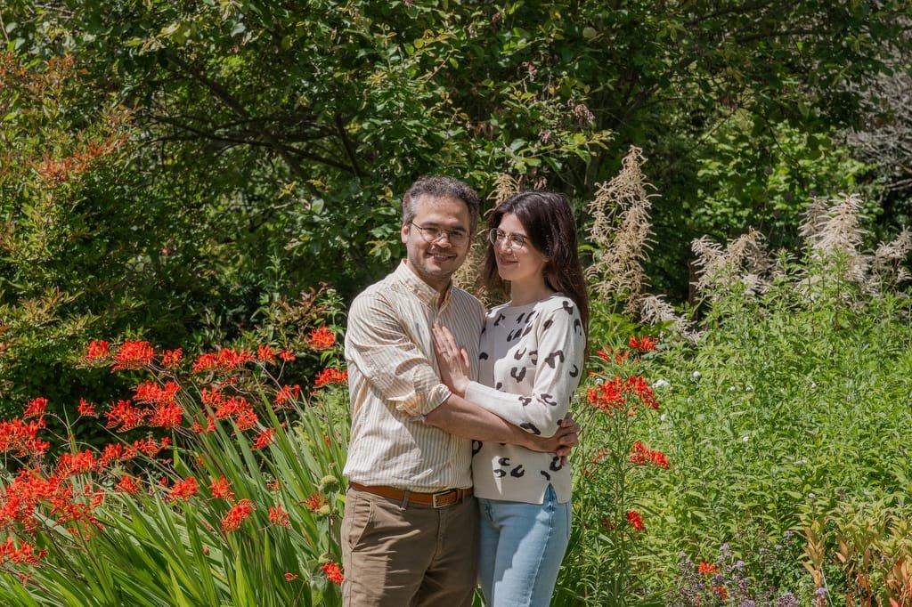 A man and a woman stand closely together in a garden, surrounded by lush greenery and red flowers. Smiling warmly, they seem to be enjoying the outdoor setting, reminiscent of a serene stroll near the Temple of Minerva Bath.