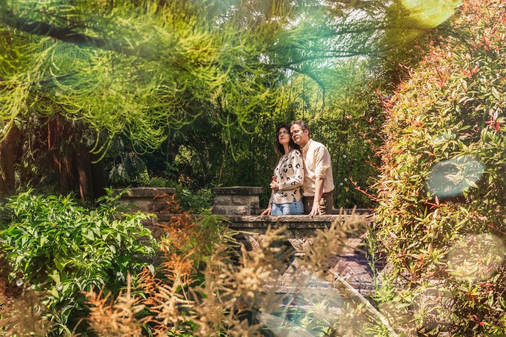 Two people stand closely together on a stone bridge in a lush garden surrounded by greenery and vibrant foliage, reminiscent of the serene ambiance near the Temple of Minerva Bath.