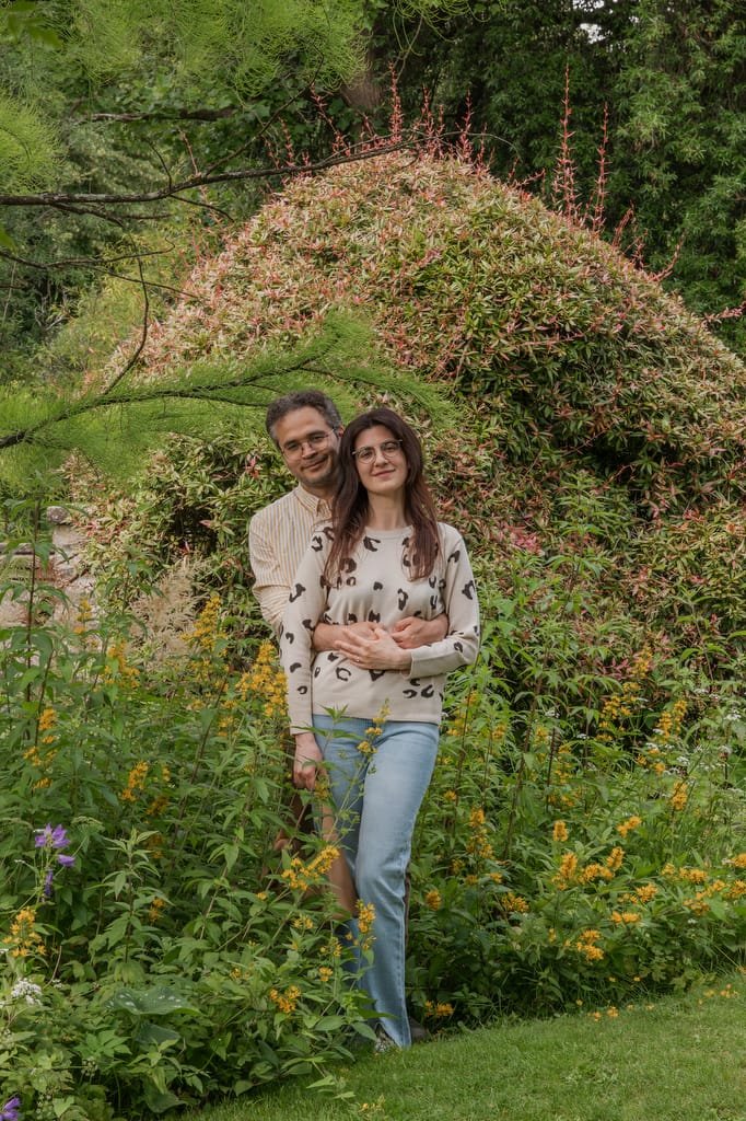 A man and woman stand close together among tall flowers and greenery, with the man embracing the woman from behind. They pose outdoors in front of a large bush and tall plants, reminiscent of a serene scene at the Temple of Minerva Bath.