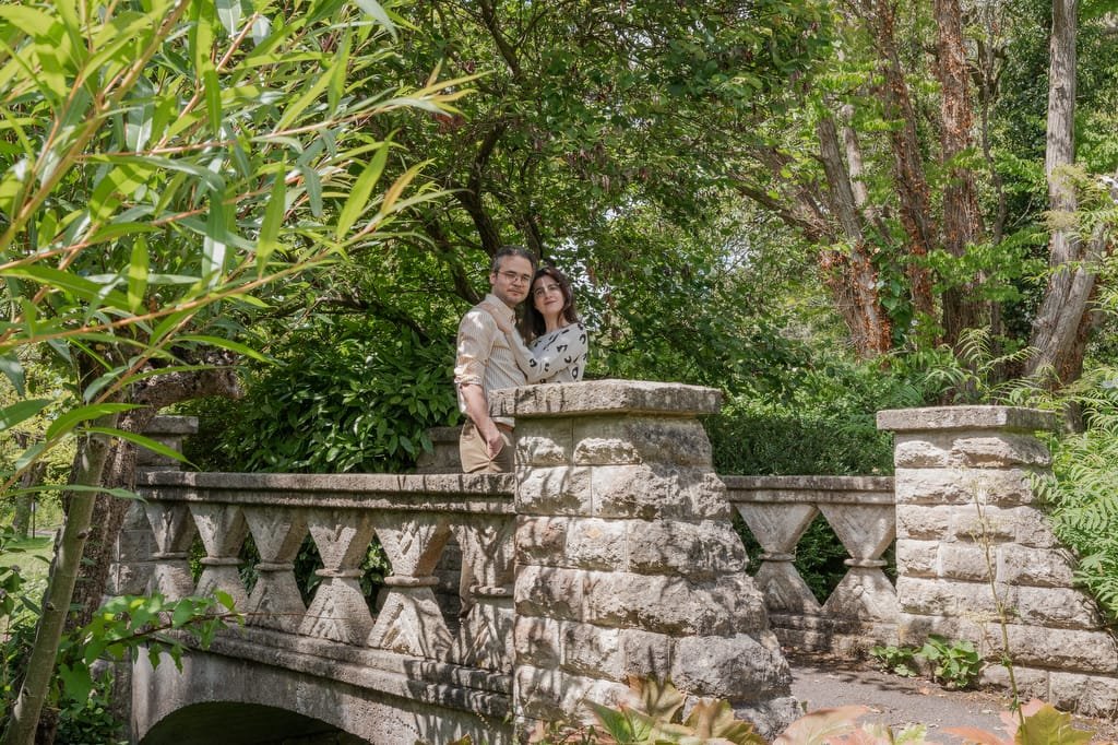 Two people stand close together on a stone bridge near the serene Temple of Minerva Bath, surrounded by lush greenery and trees.
