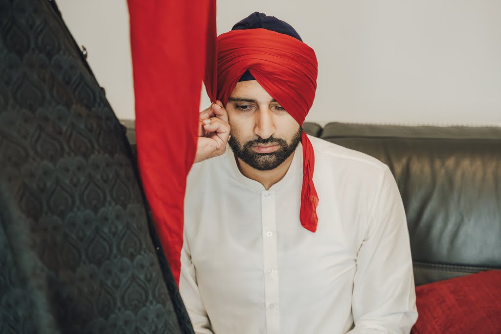 A man wearing a red turban and white traditional attire sits on a dark sofa, adjusting his left ear, embodying wedding traditions from around the world.