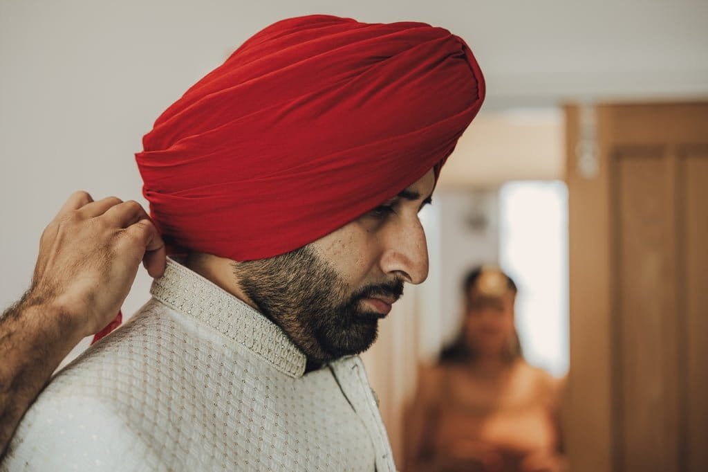In a cozy room, a man is having a red turban tied on his head by another person, embodying the rich wedding traditions from around the world. He complements this tradition with a light-colored textured jacket, ready for his special day.