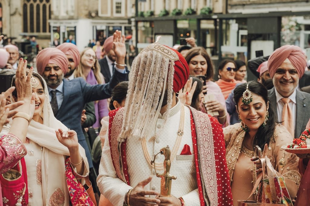 A groom in traditional Indian attire with a floral veil, surrounded by smiling guests, walks through a lively outdoor setting during a celebration, captured beautifully with Fuji Cameras for weddings.