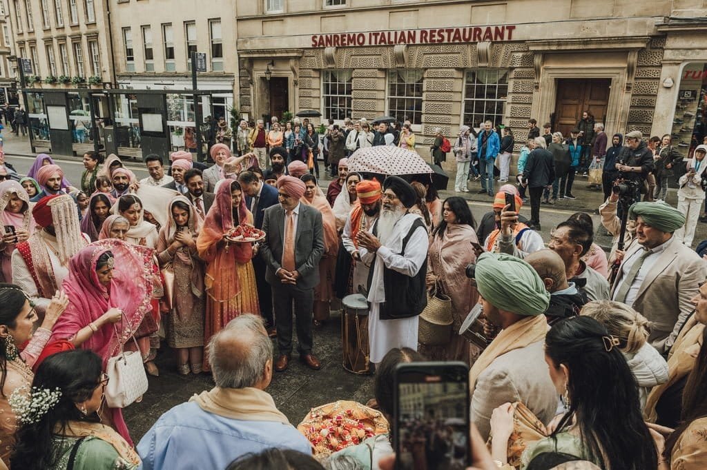 A crowd of people in traditional attire gathers outside the Sanremo Italian Restaurant, possibly to celebrate wedding traditions from around the world. Some individuals hold trays with items while others stand under umbrellas.