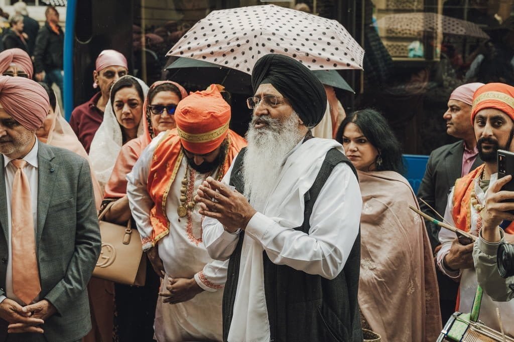 A group of people, some wearing turbans, gather outdoors. One man with a white beard and black turban appears to be leading the gathering, reminiscent of wedding traditions from around the world. An umbrella is visible in the background.