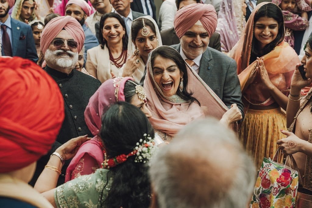A group of people, including women in traditional attire and men in stylish suits and turbans, are smiling and celebrating outdoors. Embracing wedding traditions from around the world, one woman is particularly enthusiastic, laughing and embracing others.