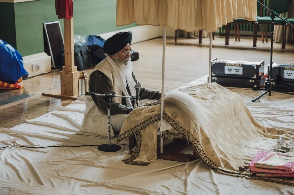 An individual wearing a turban and traditional attire sits before a draped platform, possibly involved in a religious or ceremonial act, with microphones and electronic equipment around—an evocative scene reminiscent of wedding traditions from around the world.