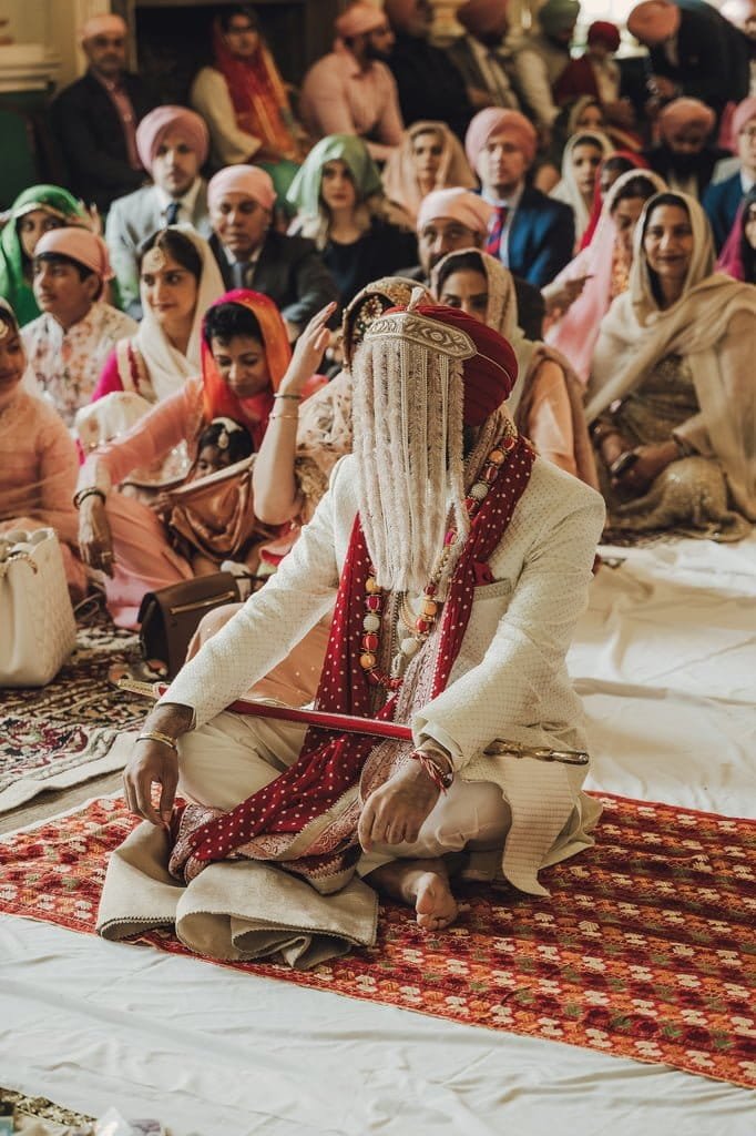 A person dressed in traditional attire with a white-covered face sits cross-legged on a patterned rug during a ceremony, embodying wedding traditions from around the world. Others in colorful clothing are seated in the background, adding to the vibrant atmosphere of cultural celebration.