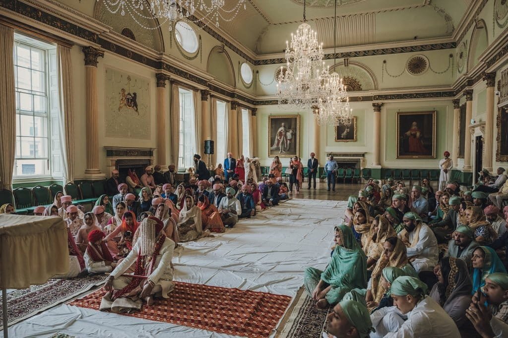 A large group of people, dressed in traditional attire, participate in a cultural or religious ceremony inside an ornate hall with chandeliers and large windows, showcasing wedding traditions from around the world.