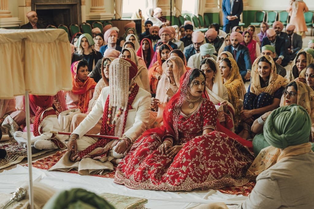 A couple in traditional attire participates in a wedding ceremony at a Sikh temple. Guests, seated on the floor and adorned in colorful clothing and head coverings, embrace the rich Wedding Traditions from around the world.
