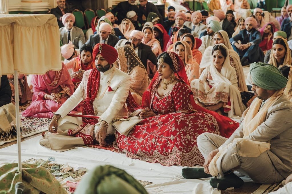 A bride and groom clad in red traditional attire sit cross-legged, surrounded by guests at a cultural ceremony inside a hall, showcasing wedding traditions from around the world.