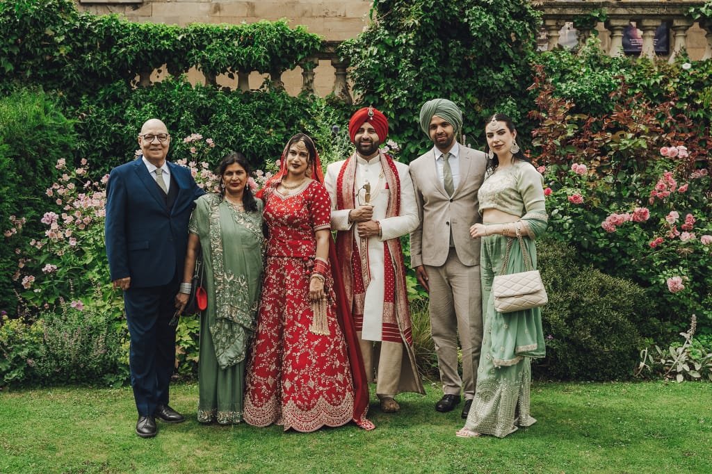 A group photo of six people, including a bride in a red wedding dress and groom in a white and red outfit with a turban, posing in a garden. The others are dressed in formal attire. Captured by the Guildhall Bath Wedding Photographer, it's a moment of timeless celebration.