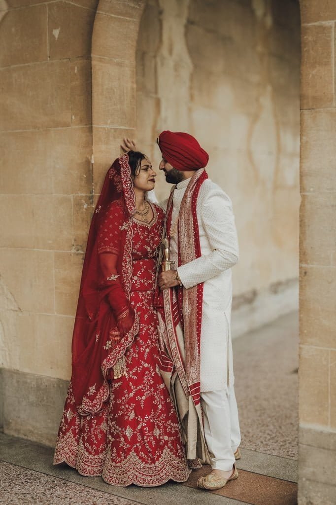 A couple dressed in traditional South Asian wedding attire stands under an archway, symbolizing wedding traditions from around the world. The woman wears a red lehenga, and the man dons a white sherwani with a red turban, exchanging amorous glances.