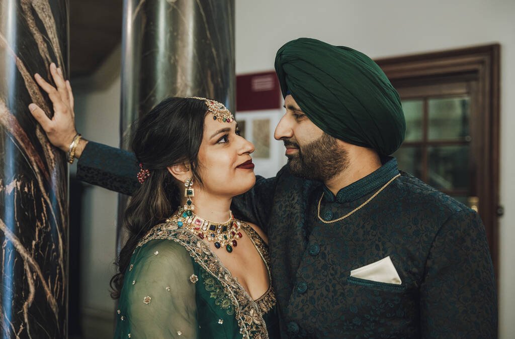 A couple dressed in traditional attire, with the woman wearing detailed jewelry and the man in a turban, stand close to each other, gazing into each other's eyes near a marble pillar—a moment beautifully captured by Fuji Cameras for weddings.